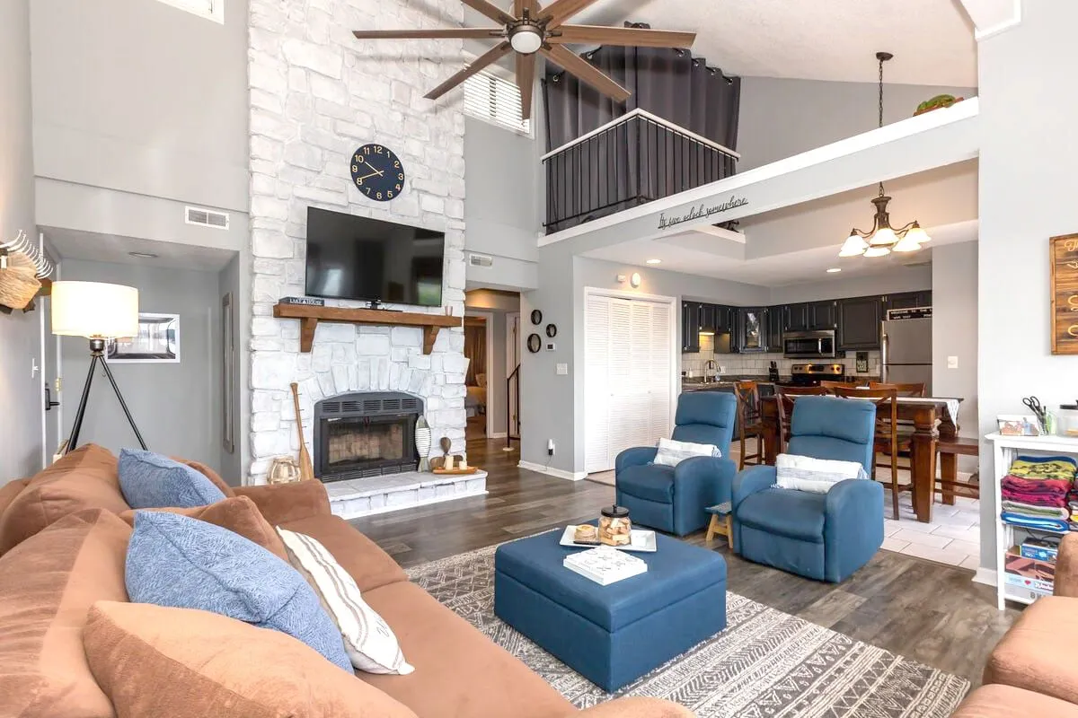A spacious open-concept living area featuring a stone fireplace, a large comfortable sofa, blue armchairs, and a view into a well-equipped kitchen. The room is warmly lit, creating a welcoming environment for guests to relax, entertain, or enjoy meals together.