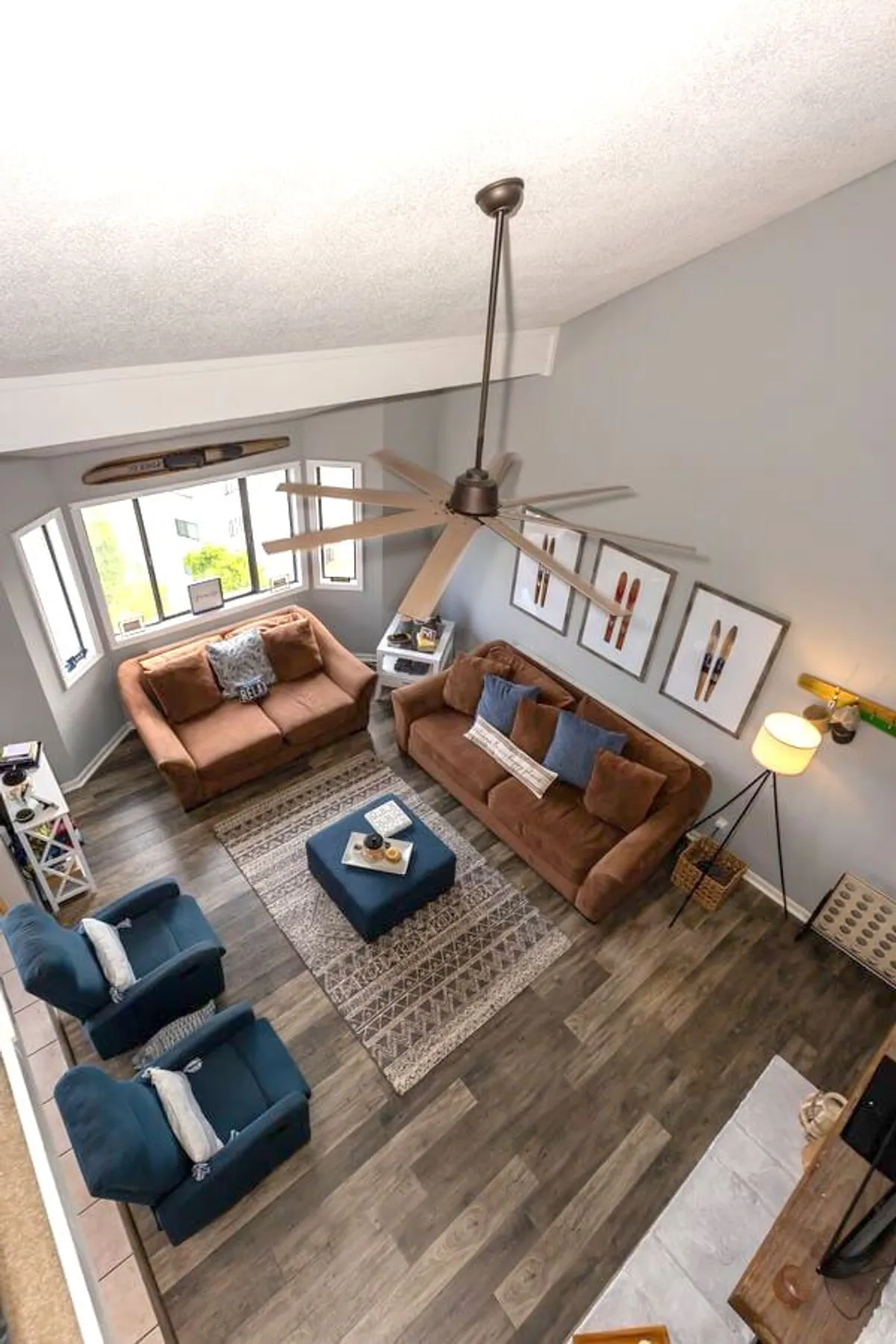 A cozy lounge area featuring brown sofas, blue accent chairs, a coffee table, and prominent wall art. The space is arranged to foster social gatherings, games, and movie nights, with a large ceiling fan providing a touch of resort-style ambiance.