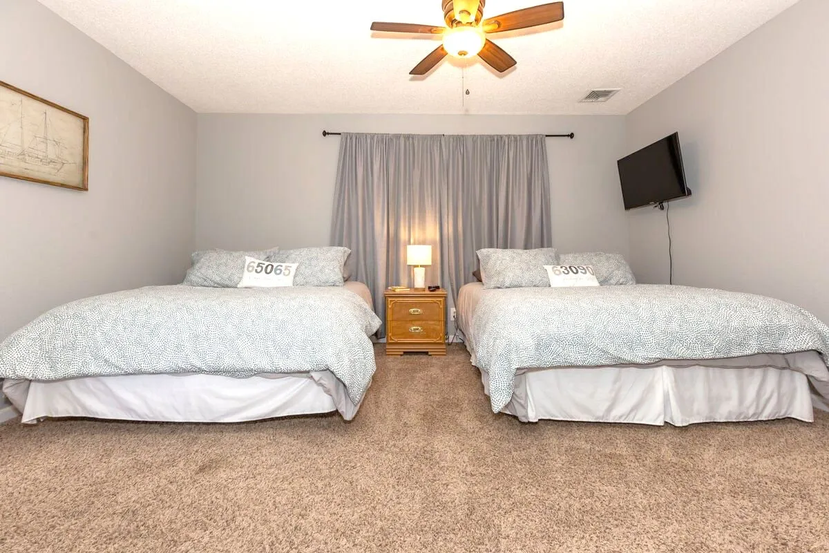 A comfortable bedroom setup with two beds, a nightstand with a lamp, wall-mounted TV, curtains, and framed artwork. The arrangement suggests flexibility for groups or families, with tranquil lighting ideal for winding down after adventures.