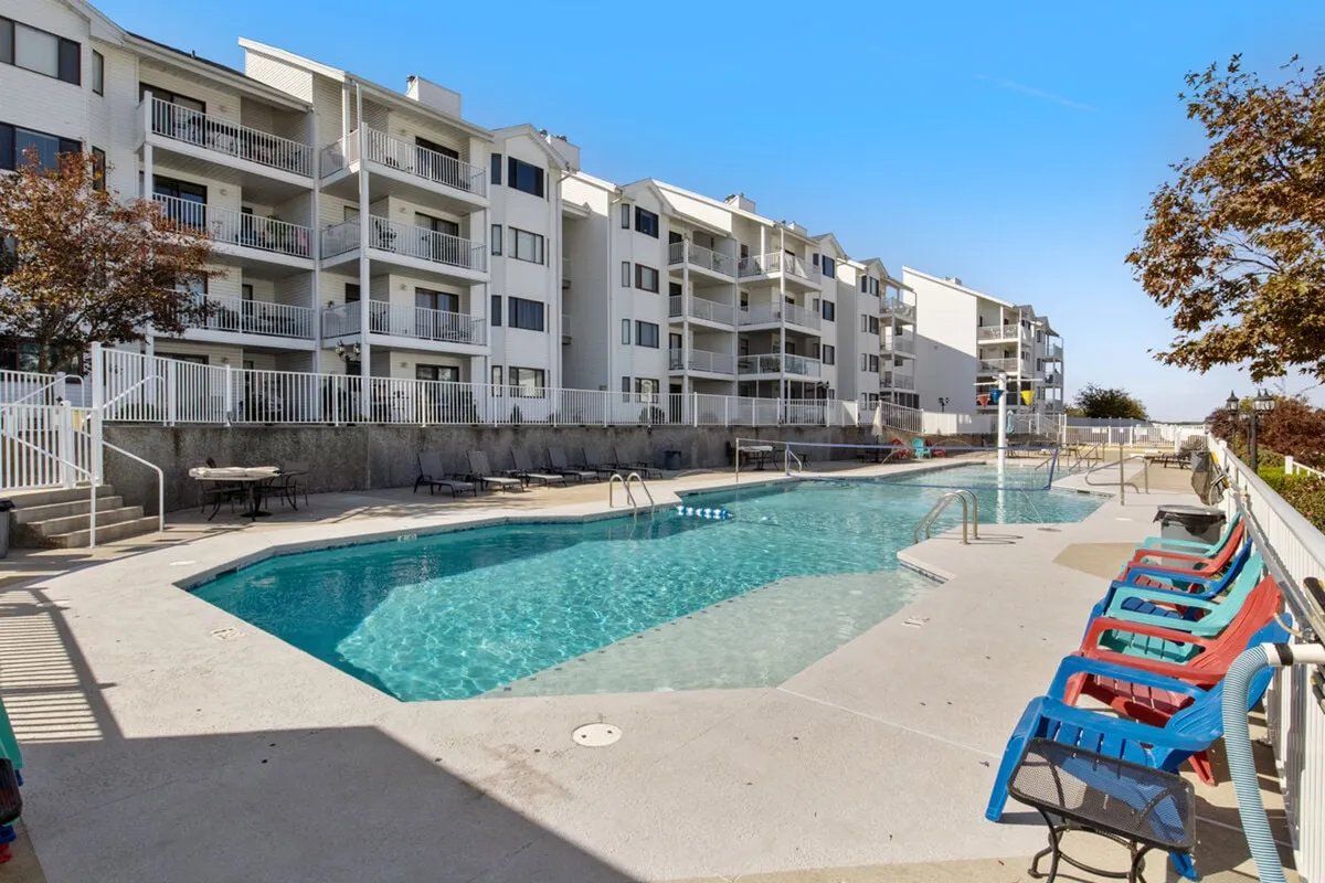 A sunny outdoor pool area with inviting blue water, sun loungers, and the apartment balconies visible in the background. The scene communicates relaxation and leisure in a well-maintained communal setting.