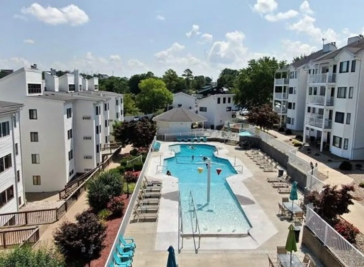 A sunlit outdoor swimming pool is surrounded by lush greenery, comfortable lounge chairs, and neighboring buildings. The inviting water glistens, while the area exudes a relaxed resort-like vibe perfect for both children and adults to enjoy together.