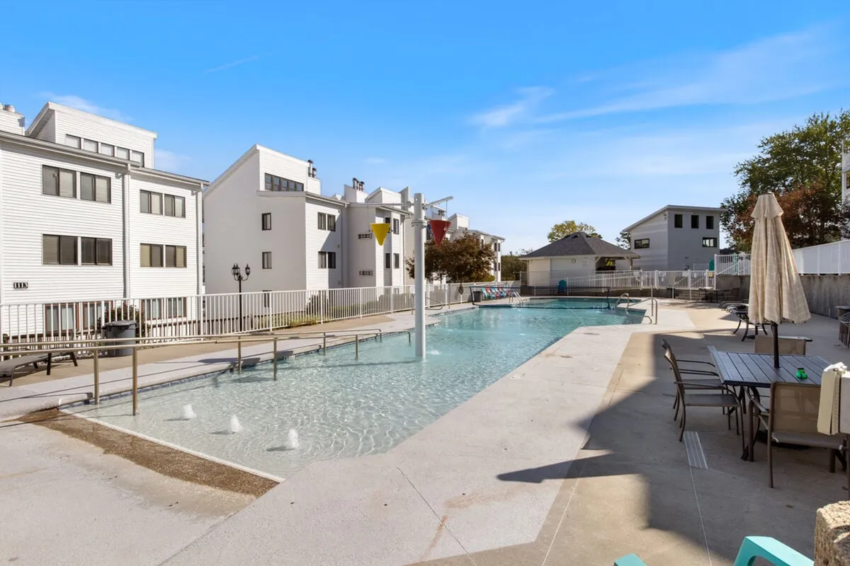 A cheerful scene featuring a large swimming pool with outdoor furniture, fountains, and surrounding buildings. The space is set up for both relaxation and social gatherings, capturing the leisurely and family-friendly atmosphere of the amenities available.