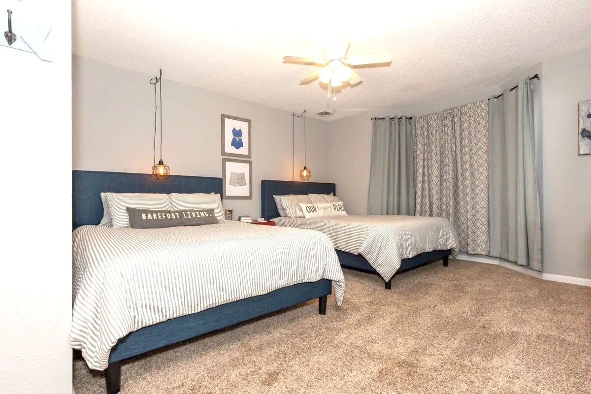 A cozy and welcoming bedroom with two twin beds featuring blue upholstered headboards and striped bedding. Decorative wall art and hanging light fixtures add a playful, family-friendly touch to the space.