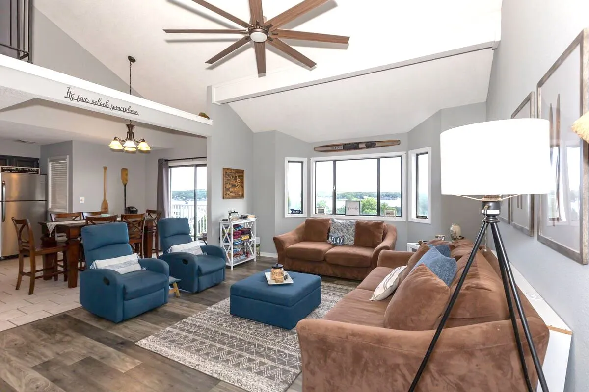 A spacious open-concept living and dining area featuring a comfortable sofa, large dining table, and expansive windows that showcase beautiful sunset views over the lake. The space is bathed in natural light and is accented by a ceiling fan, fostering an inviting and relaxed atmosphere ideal for gathering.