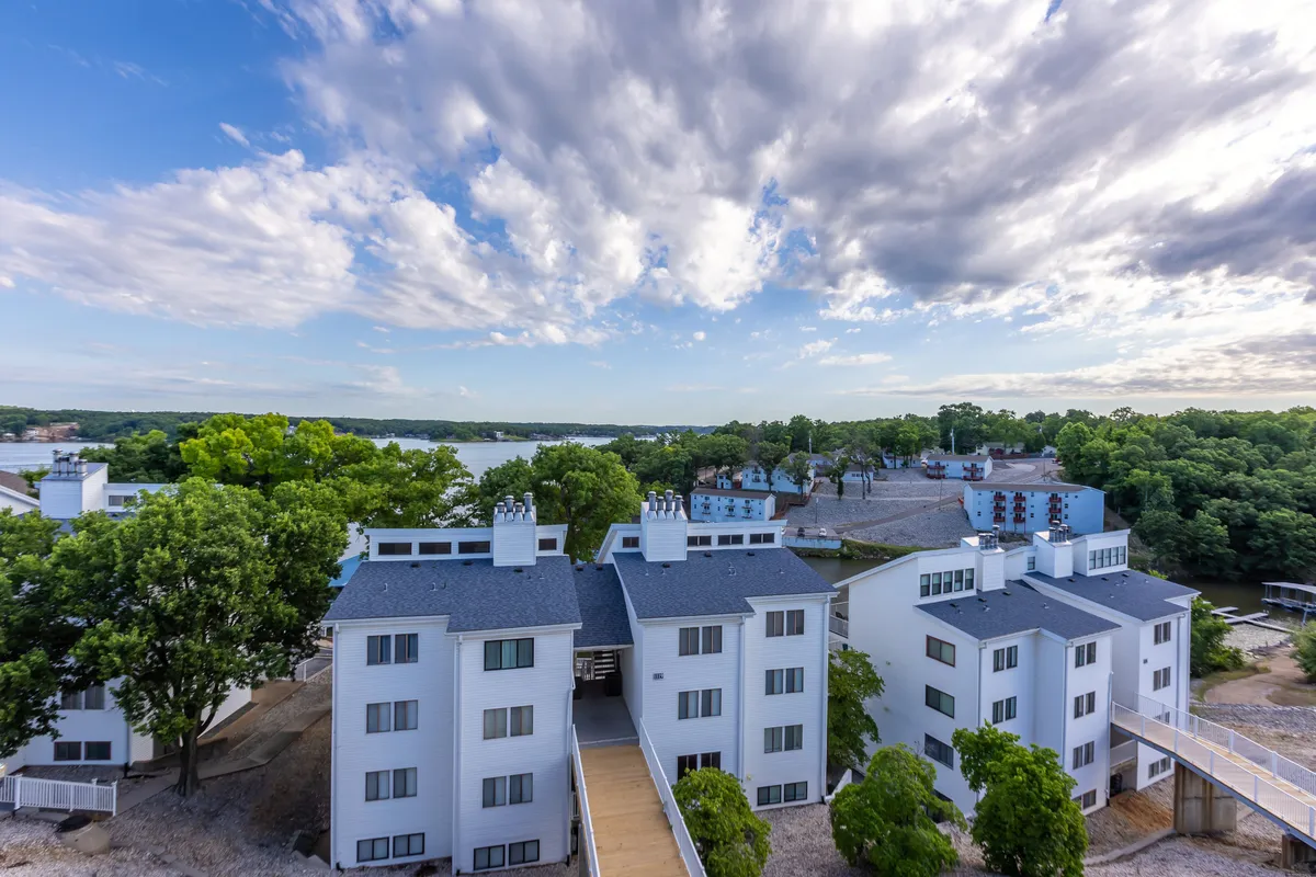 This exterior shot showcases white multi-story buildings nestled among lush green trees, with sparkling lake water and a vast sky in the background. The setting is peaceful and inviting, suggesting opportunities for boating, fishing, nature walks, and lakeside relaxation.