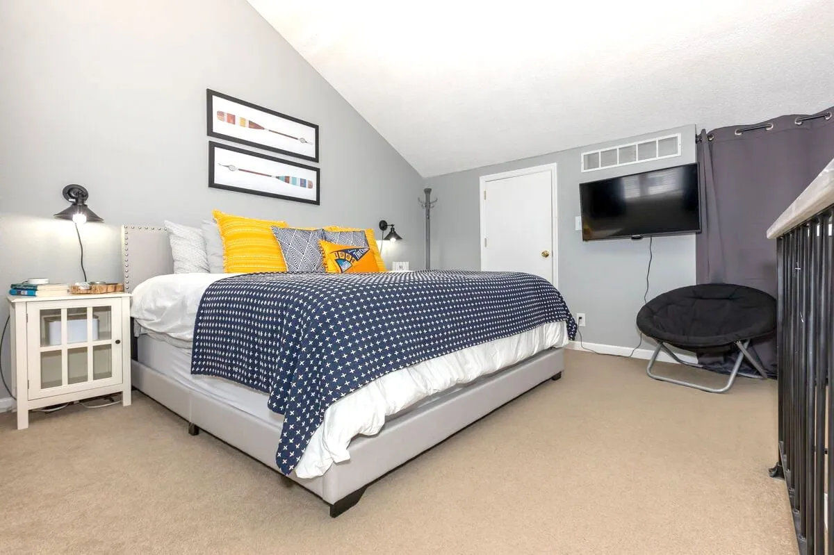 A stylish bedroom featuring a cozy bed with patterned bedding, decorative pillows, artwork on the wall, a nightstand with a lamp, and a television. The space is filled with soft light, inviting relaxation and comfort. The television and ample bed surface underline family-friendly flexibility.