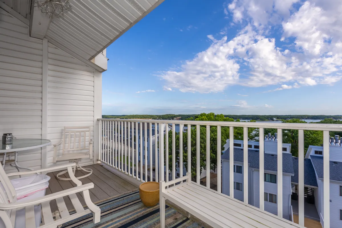 A private balcony with a railing overlooks a picturesque landscape, offering comfortable seating and a decorative pot. The view suggests a tranquil lakeside setting, with perfect spots for morning coffee or evening relaxation as the sun rises or sets.